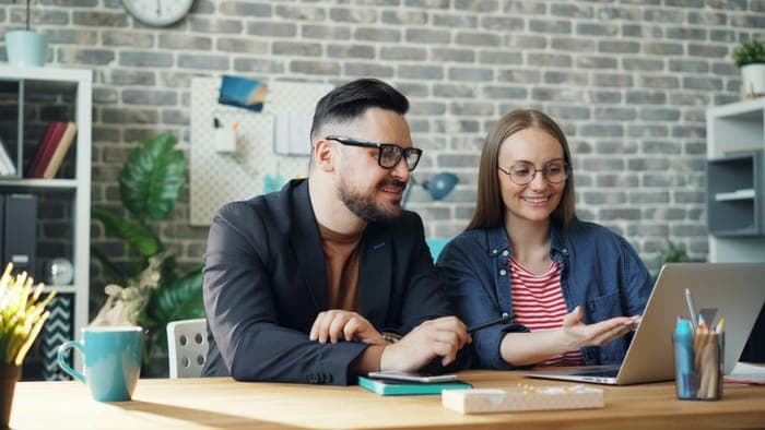 Two colleagues smiling at a laptop in an office with a brick wall